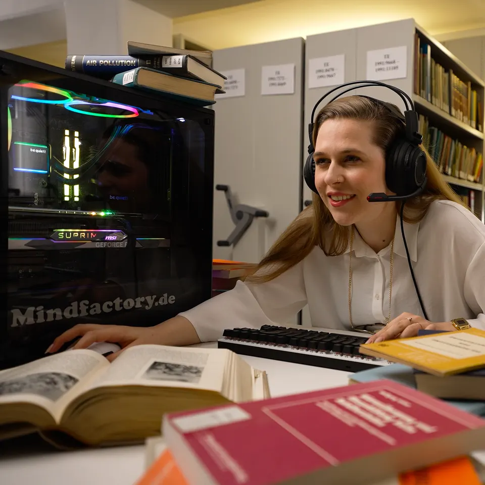 Frau mit Headset an einem Gaming-PC zwischen Bücherregalen, Kampagne für Mindfactory